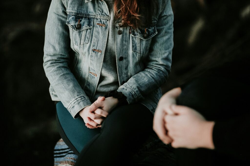 An image representing what actually happens in a therapy session. Woman sitting wearing grey jacket facing another woman, both with their hands clasped in a comfortable position. 