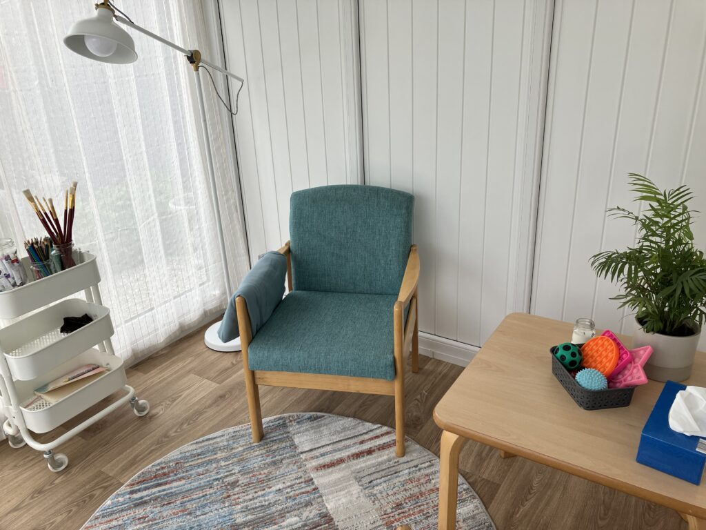 A photo showing neurodivergent Therapy in Stirling therapy room. A photo of an art trolley, standing lamp, turquoise chair, striped rug and a table with a plant, fidget toys and tissues.