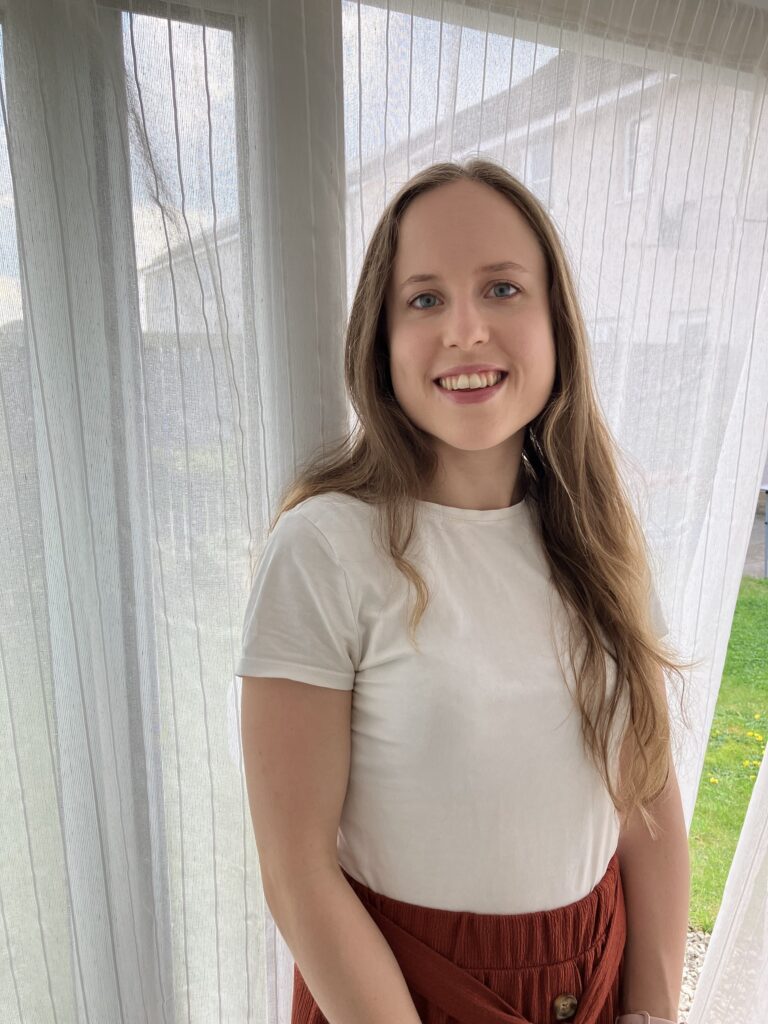 A photo of Caitlin smiling standing in the open doorway of the neurodivergent therapy in Stirling room. Caitlin is wearing a white top and brown skirt.