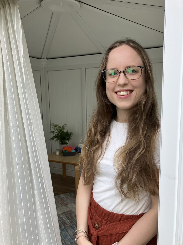 A photo of Caitlin from Caitlin Imray neurodivergent therapy in Stirling smiling wearing glasses, a white top and a brown skirt. She is standing in the doorway of her therapy space.