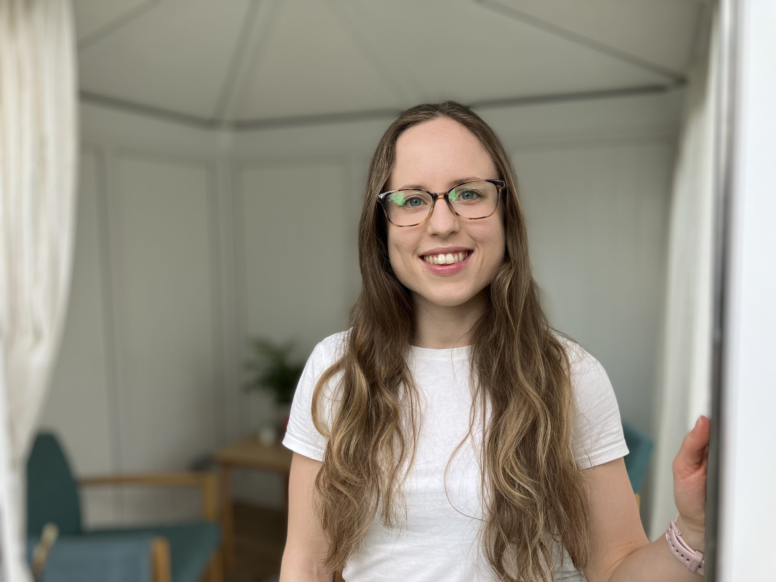 A photo of Caitlin wearing glasses and a white top standing in the doorway of her therapy space.