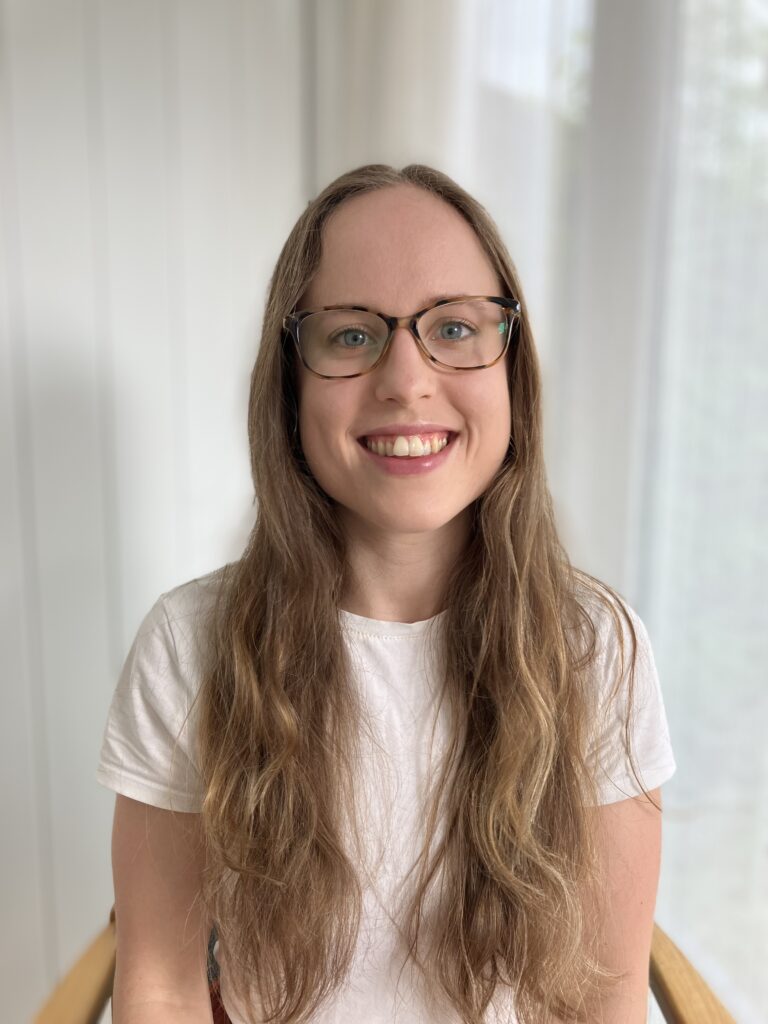 A photo of Caitlin smiling sitting by a window and wearing glasses and a white top in her neurodivergent therapy in Stirling room.