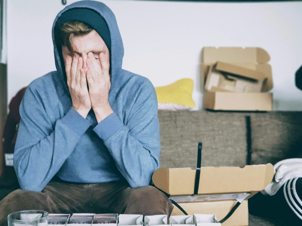 A person wearing a blue hooded sweatshirt with their hands over their face and piles of boxes around them. They look emotionally dysregulated. Image represents blog post title 'is therapy effective for helping autistic people with emotional regulation'